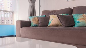 A modern living room with a brown sofa, teal and brown patterned cushions, a window with a view of buildings, and a blue wall. - Apartamento no Guaruja com Vista para o Mar (Guarujá)