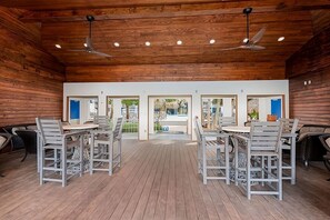 A spacious indoor area with wooden walls and ceiling, featuring multiple tables and chairs, and a view into an outdoor area through glass doors.