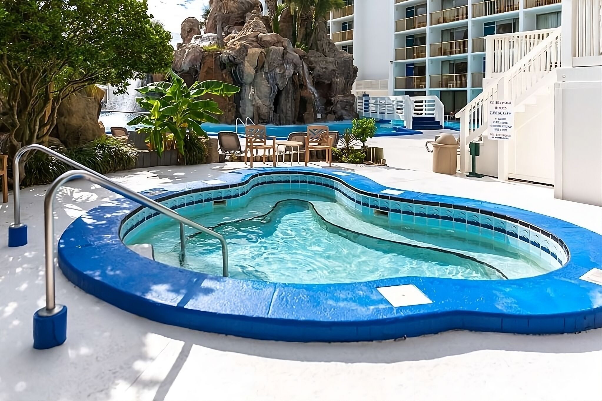 A hot tub surrounded by a pool, with a rocky backdrop and a building in the background.