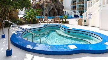 A hot tub surrounded by a pool, with a rocky backdrop and a building in the background.