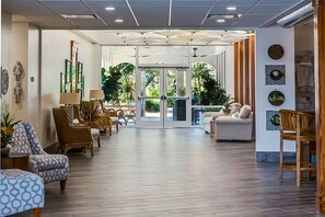A spacious lobby with a modern design, featuring a central glass door leading to an outdoor area, surrounded by comfortable seating and decorative wall art.