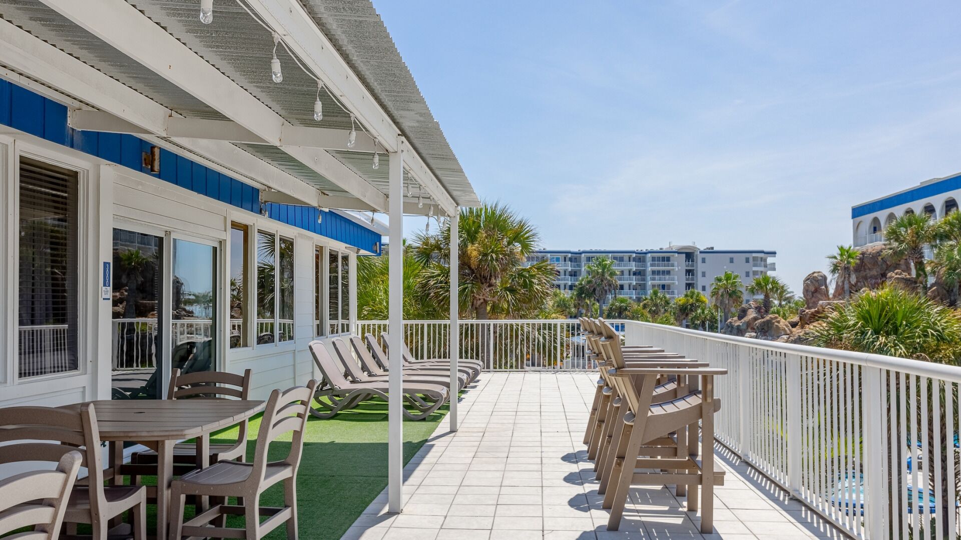 Een hotelbalkon met buitenmeubilair en uitzicht op de oceaan, met op de achtergrond een gebouw met blauwe accenten.