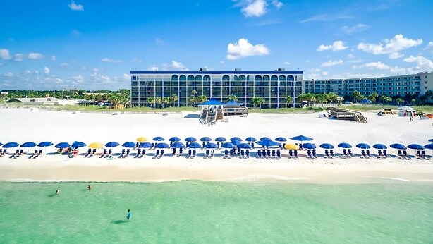 Un complexe hôtelier de bord de mer doté d’un grand bâtiment, d’une eau turquoise et limpide, et de nombreux parasols.