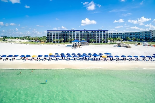 A beach resort with a large building, clear turquoise water, and numerous umbrellas.