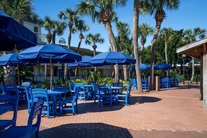 An outdoor patio area with blue plastic chairs and umbrellas, surrounded by palm trees. - Peaceful Retreat for Your Beach Escape | Outdoor Pool & Garden Courtyard (Fort Walton Beach)