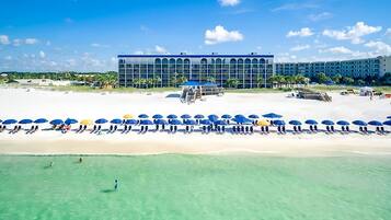 A beachfront hotel with a row of beach umbrellas and lounge chairs, a clear view of the ocean, and a large building with a modern design.