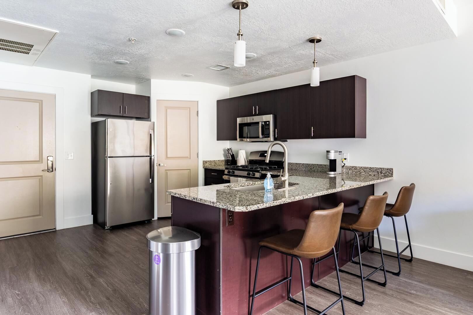 A modern kitchen with dark cabinets, stainless steel appliances, a granite countertop, and bar stools.