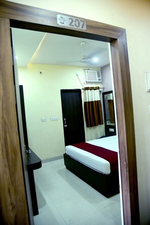 Quarto de hotel com cama, escrivaninha, espelho, janela com cortinas e ventilador de teto.