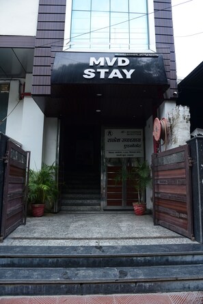 Entrada de hotel com uma placa indicando 'MVD STAY' e um número de contato. Há duas plantas em vasos na entrada.