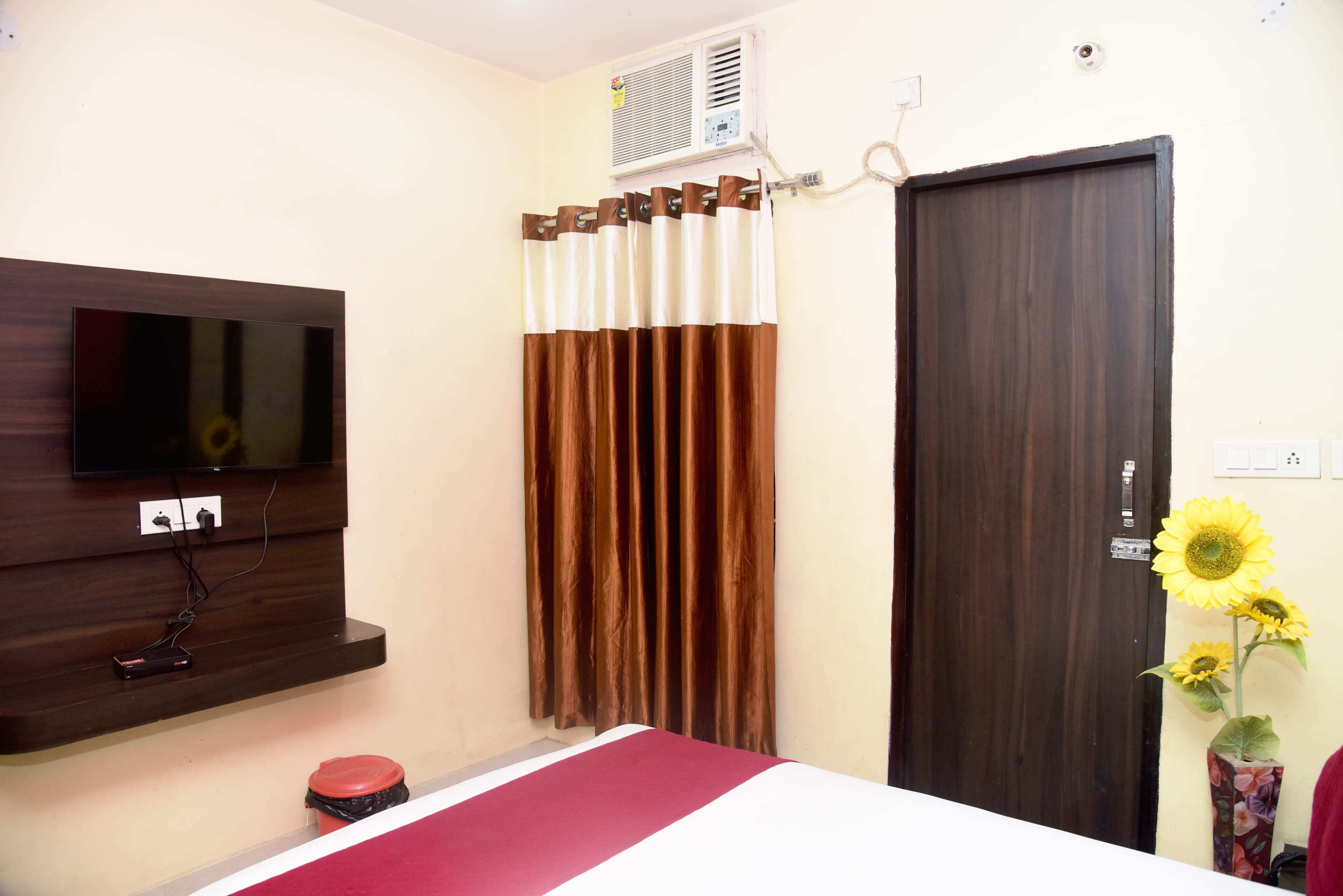Habitación de hotel con televisor de pantalla plana montado en la pared, una puerta de madera oscura, una ventana con cortinas marrones y un pequeño cesto de basura.