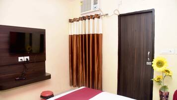 Habitación de hotel con televisor de pantalla plana montado en la pared, una puerta de madera oscura, una ventana con cortinas marrones y un pequeño cesto de basura.