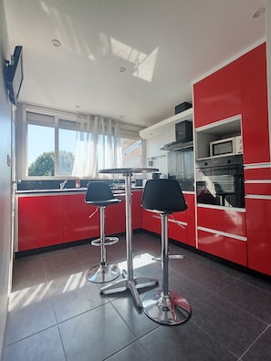 A modern kitchen with red cabinetry, two bar stools, a window with curtains, and a microwave on the countertop. - JULIEN'S 4-ROOM APARTMENT (Tarbes)