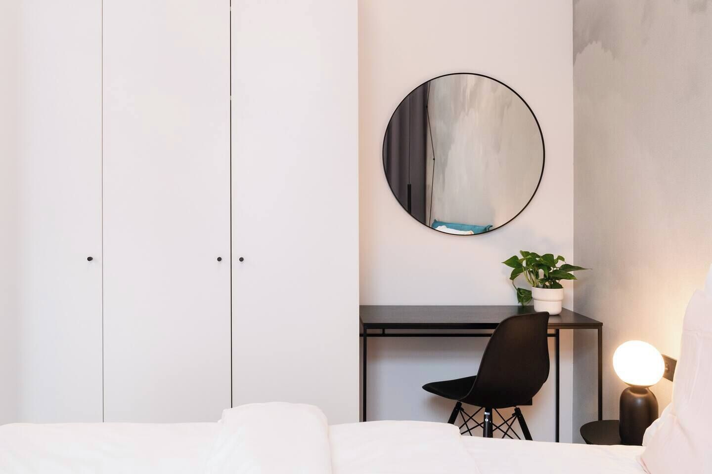 A bedroom with a bed, a desk, a chair, a round mirror, and a bedside lamp.