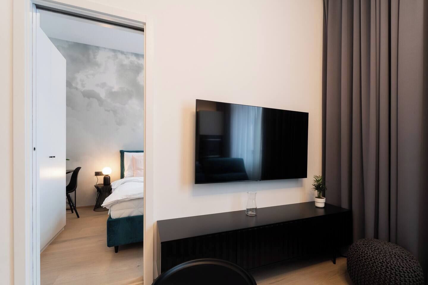 Une chambre d’hôtel moderne, dotée d’un grand téléviseur à écran plat fixé au mur, d’une console sombre, d’un lit avec une literie blanche et d’une petite plante en pot.