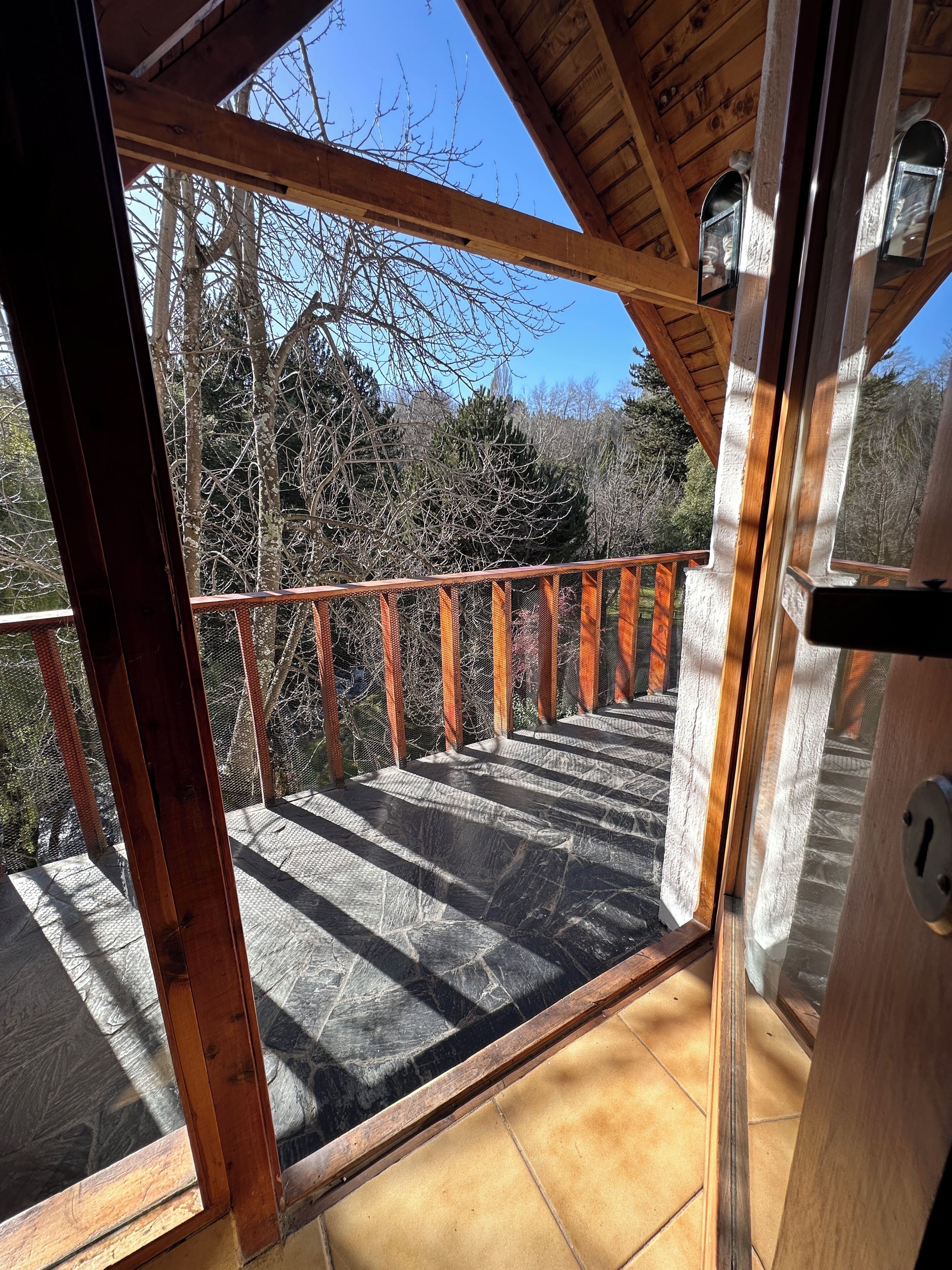 A wooden balcony with a metal railing, a stone floor, and a view of trees and sky.