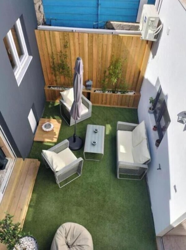 A small patio area with a wooden fence, a white wall, and a green artificial turf. There are two white chairs, a small table, and a potted plant. - The Violet House (Costa Nova)