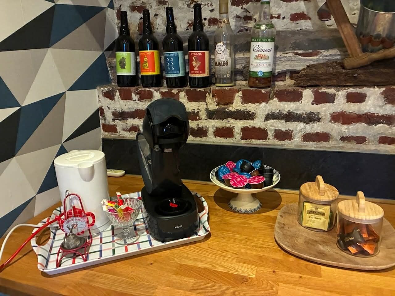A coffee machine on a tray with a mug, a sugar bowl, and a plate of cookies. In the background, there are bottles of wine and a brick wall.