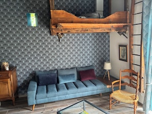 A room with a patterned wallpaper, a blue sofa with cushions, a wooden ladder, and a chair with a woven seat. - Renovated Old Norman Stable (Luneray)