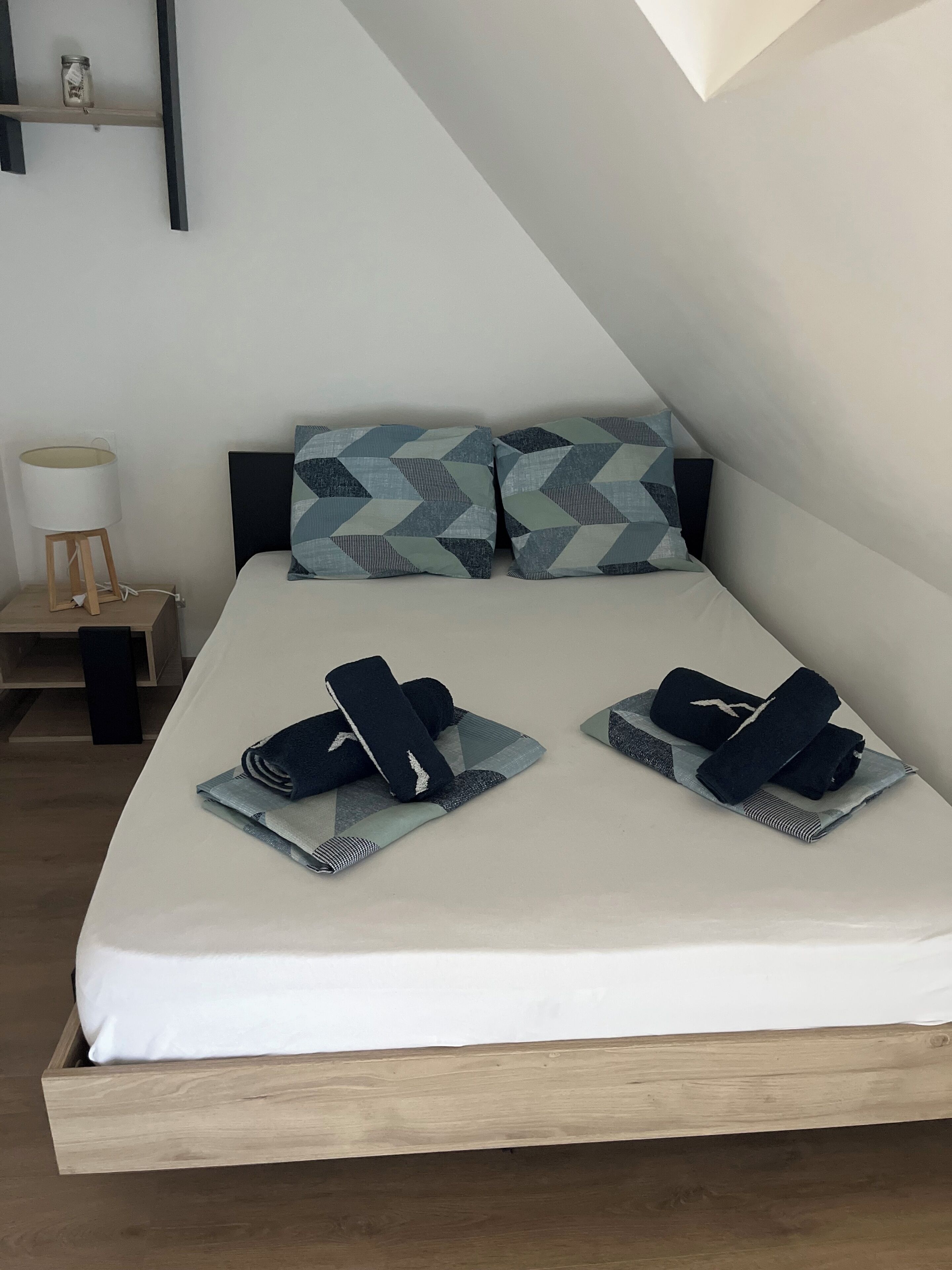Una cama bien hecha con almohadas y toallas dobladas, una mesita de noche con una lámpara y una estantería con un frasco.
