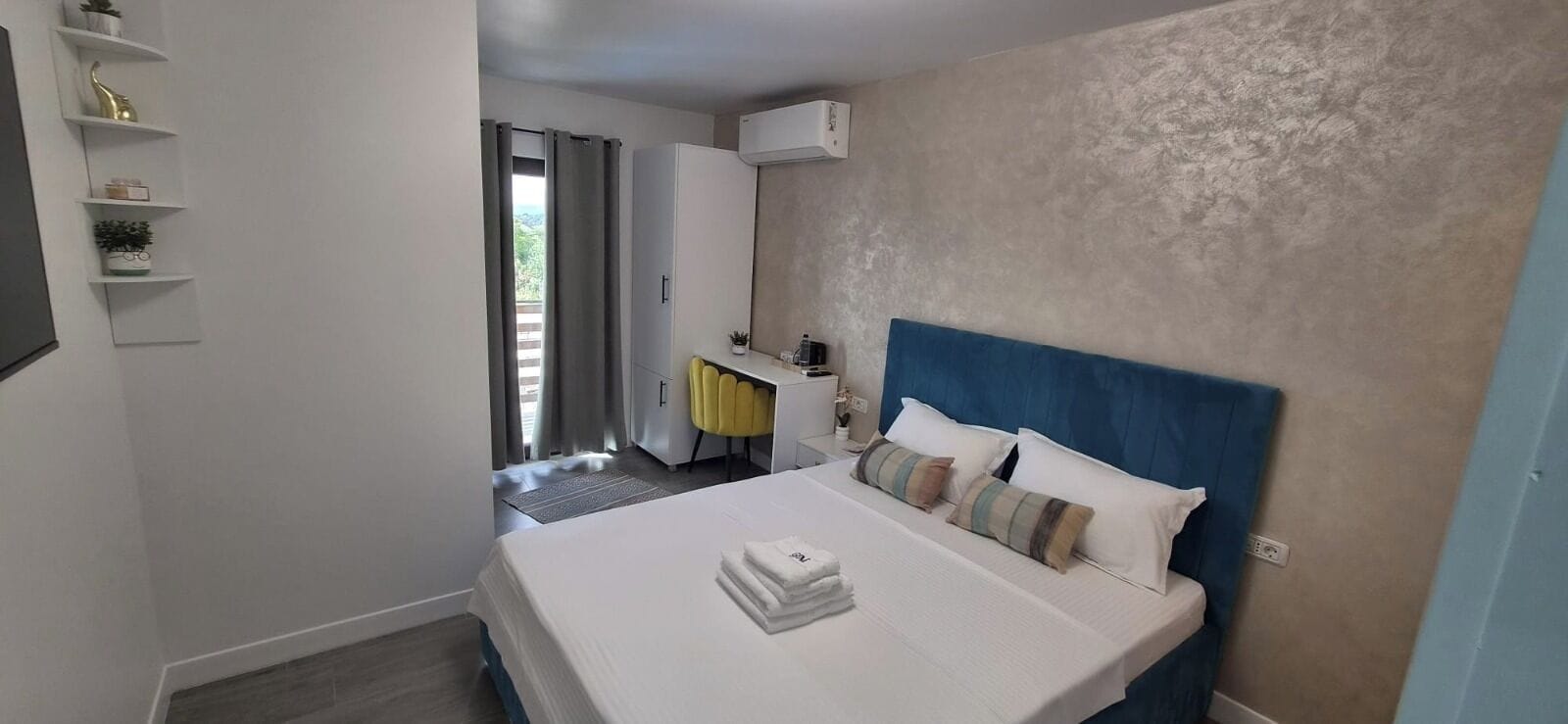 A hotel room with a bed, a bedside table, a nightstand, a wardrobe, a window with curtains, and a wall-mounted air conditioning unit.