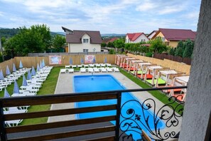 A pool area with lounge chairs and umbrellas, surrounded by a wooden fence, with houses in the background. - Good Mood Pool & Smart Rooms (Ciurea)