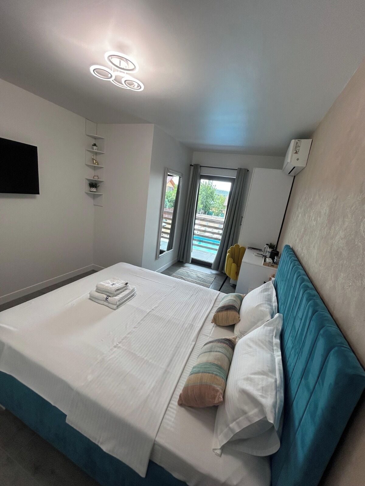 A hotel room with a bed, a flat-screen TV, a ceiling light fixture, a window with curtains, and a view of the outdoors.