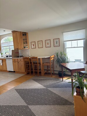 A kitchen with wooden cabinets, a dishwasher, and a dining area with a table and chairs. There are framed pictures on the wall and a large window with blinds.