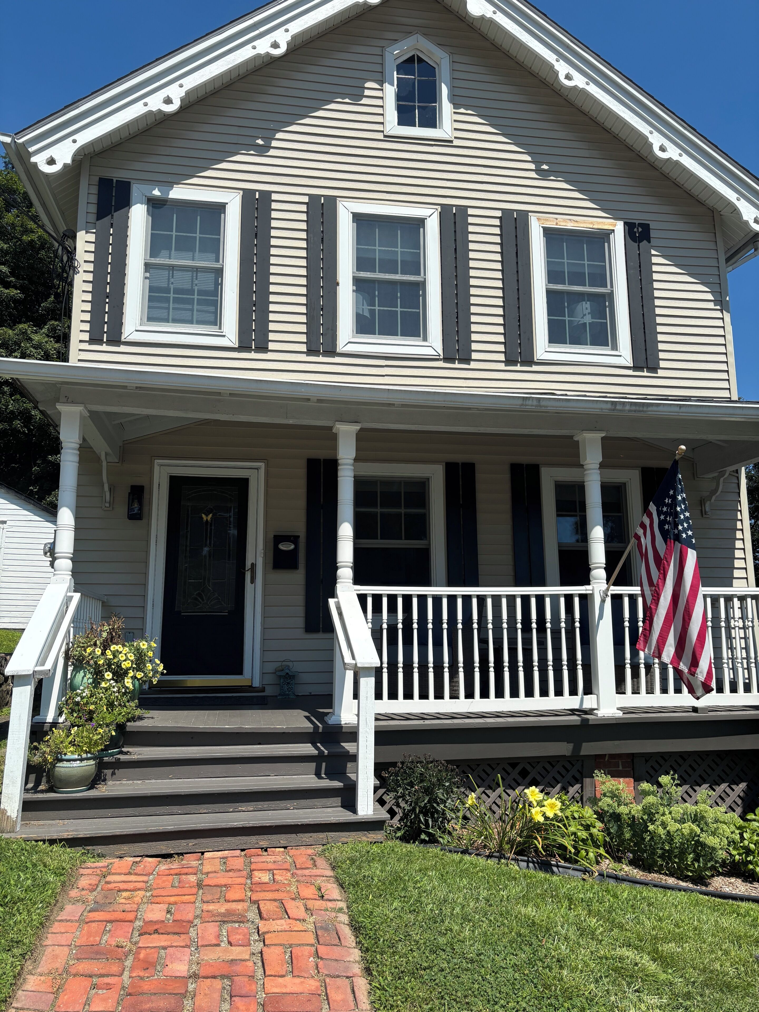 A two-story house with a front porch, a brick pathway leading to the door, and an American flag on the railing.