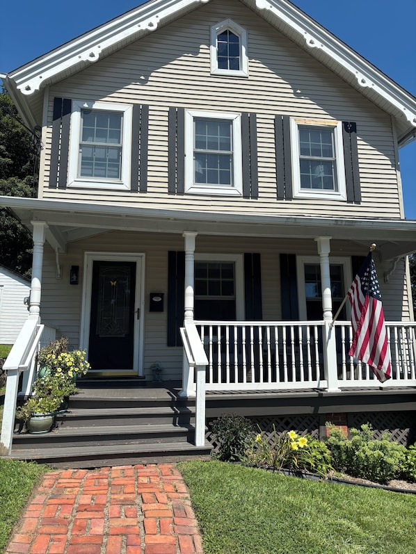 A two-story house with a front porch, a brick pathway leading to the door, and an American flag on the railing.