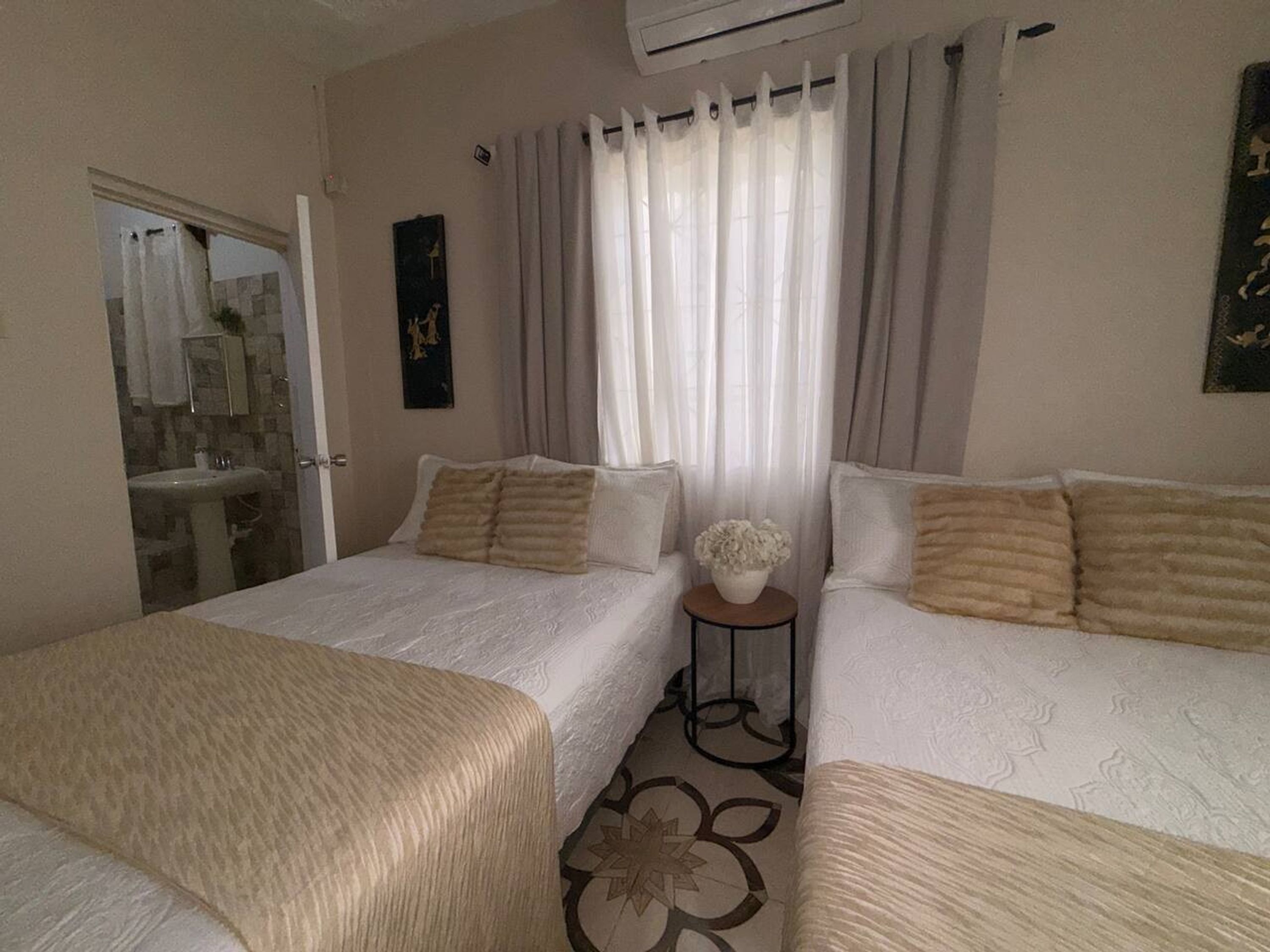 A hotel room with two beds, a small table, a bathroom with a sink, and a window with curtains.