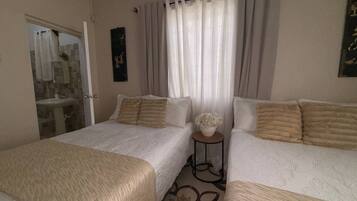A hotel room with two beds, a small table, a bathroom with a sink, and a window with curtains.