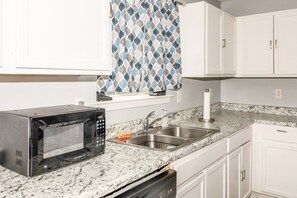 A kitchen with white cabinets, a granite countertop, a built-in microwave, a stainless steel sink, and a window with patterned curtains. - Advance Home - McDonough (McDonough)
