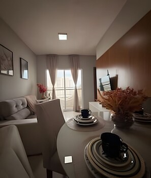 A modern living room with a sofa, a dining table with plates and a vase, and a view of a cityscape through large windows. - Apto I Piscina I Wi-Fi I Conforto- Lazer (Cabedelo)
