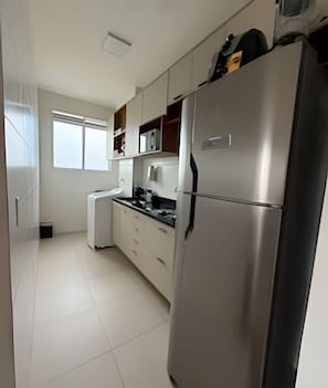 A modern kitchen with a stainless steel refrigerator, white cabinets, and a window providing natural light. - Apto I Piscina I Wi-Fi I Conforto- Lazer (Cabedelo)