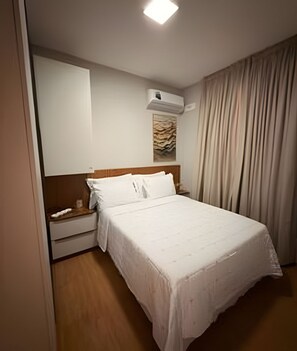 Una cama bien hecha con sábanas blancas, cabecera de madera, mesita de noche con lámpara y aire acondicionado instalado en la pared.