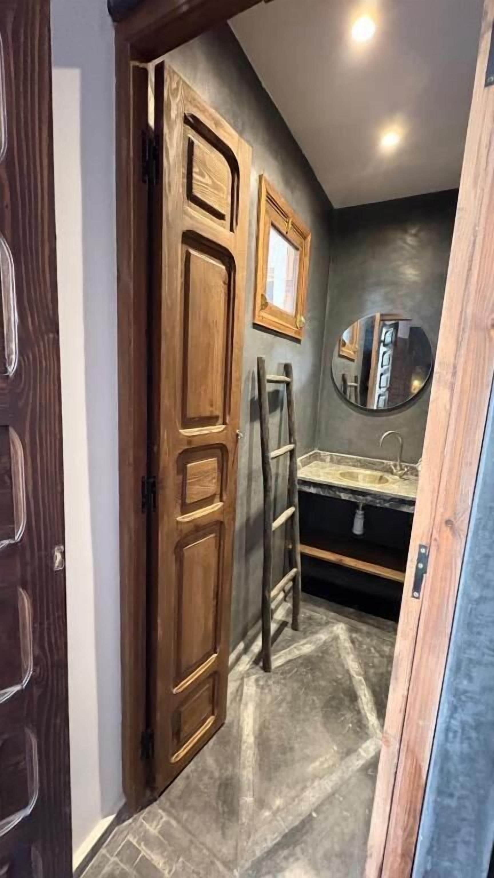 Un pasillo estrecho con una puerta de madera, una escalera y un baño con lavabo y espejo.