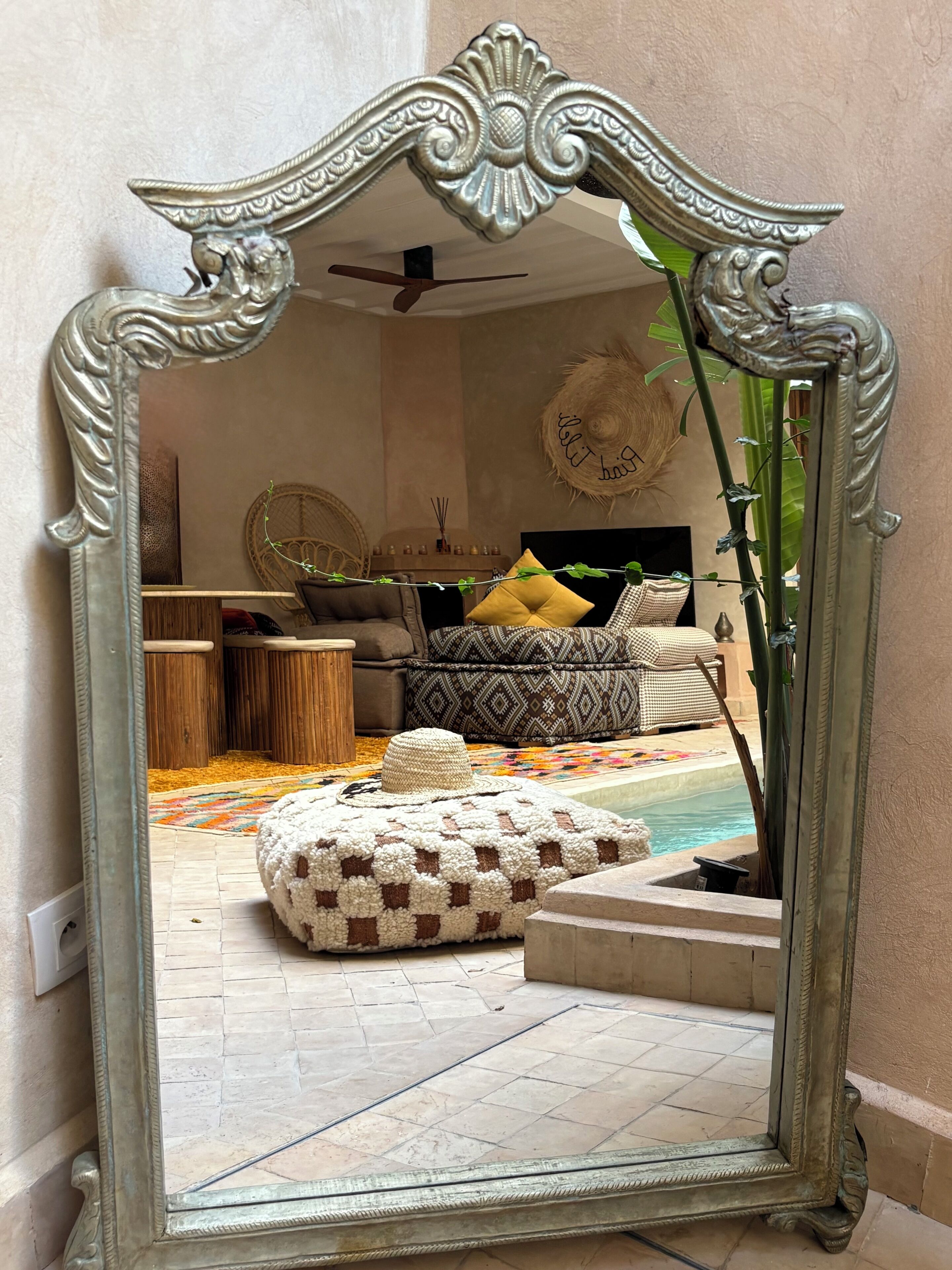 Un espejo refleja una habitación con un pouf estampado, un sombrero tejido y una cama con almohadones decorados.