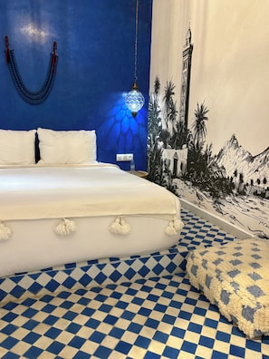 A bed with white bedding, a blue and white checkered floor, a wall-mounted light fixture, and a mural of a mountain landscape. - RIAD TILÉLI (Marrakech)