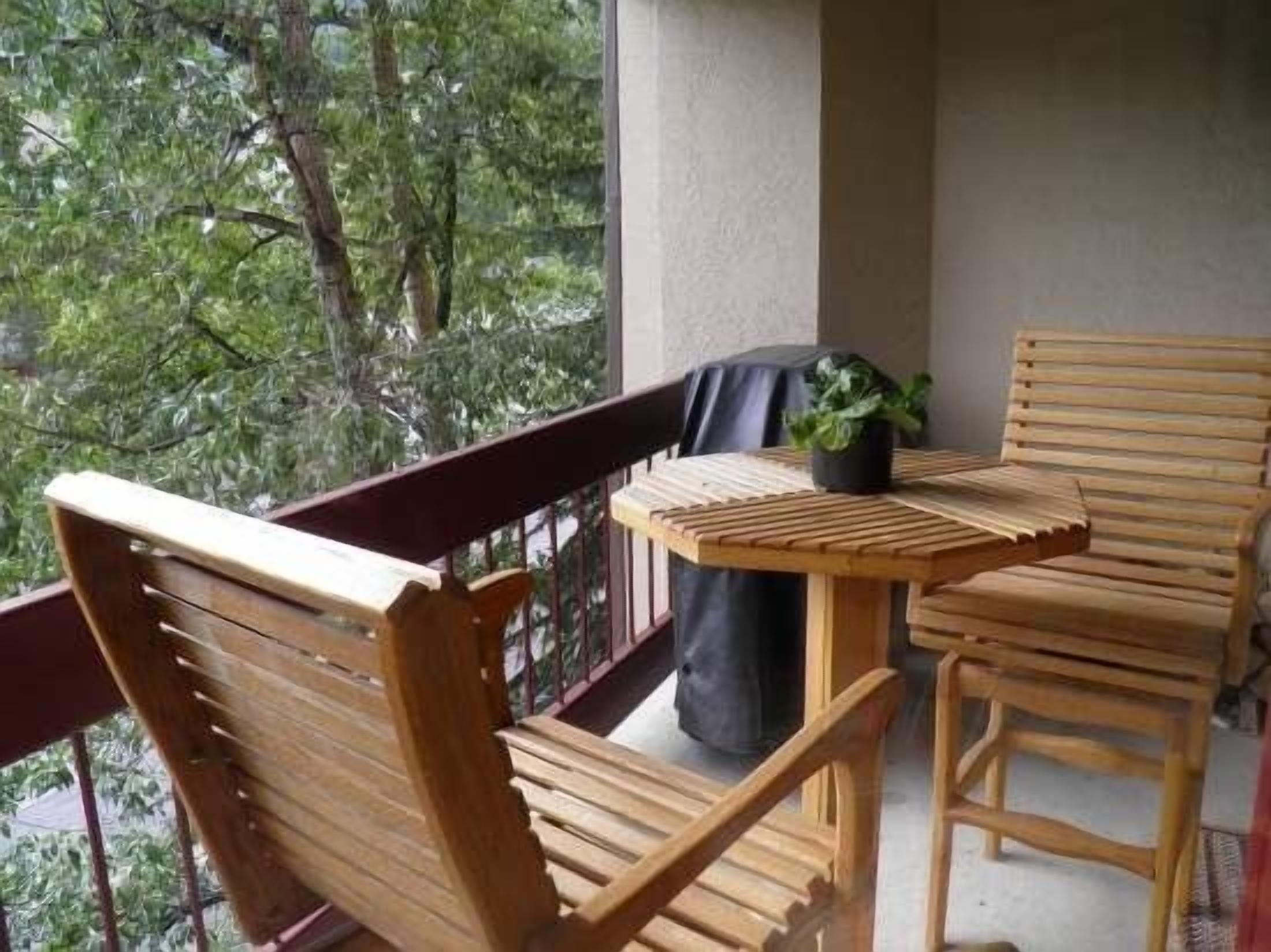 Un balcón de madera con una mesa, sillas, una planta en maceta y vista a árboles.