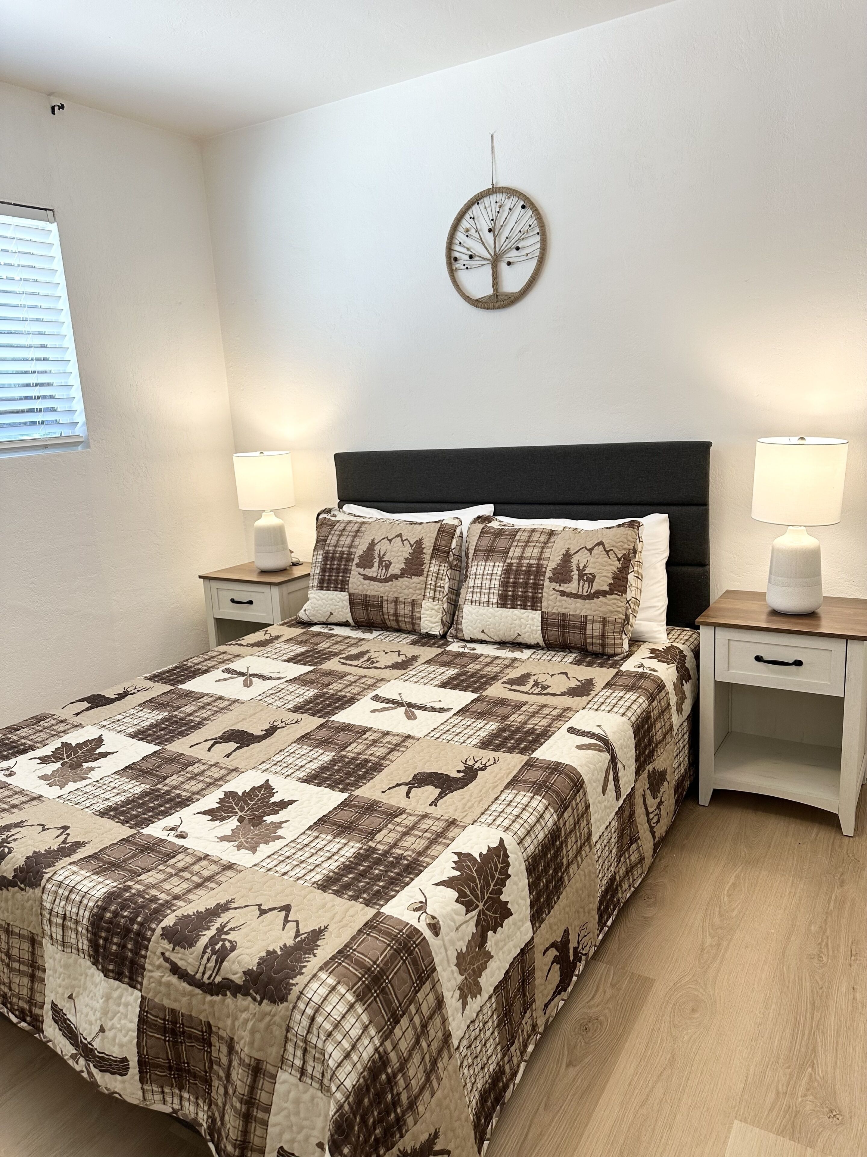 A bedroom with a bed, two bedside tables, a wall clock, and a window with blinds.
