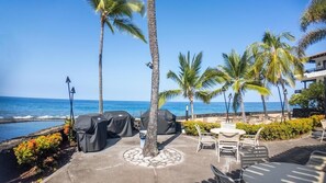 An outdoor seating area with a view of the ocean, covered barbecue grills, and palm trees. - Big Island Casa De Emdeko B326 1 Bedroom Apts (Kailua-Kona)