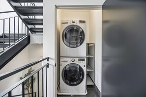 A laundry room with two stacked washing machines, a staircase, and a plant. - The Crawford HTX: Houston luxury with style, rooftop views, & local flair. (Houston)