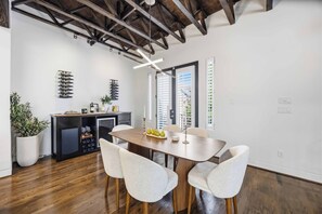 A modern dining area with a wooden table and four chairs, a kitchen with wine racks, and a large window with blinds. - The Crawford HTX: Houston luxury with style, rooftop views, & local flair. (Houston)