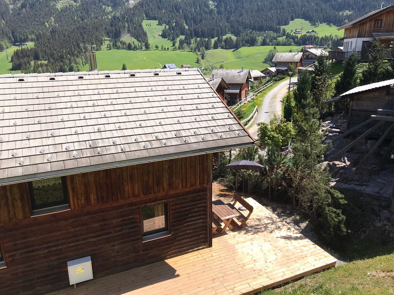 Una cabaña de madera con techo inclinado, una terraza con mobiliario exterior y vista a un paisaje rural con casas y árboles.