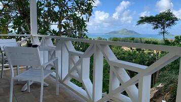A terrace with white chairs and a white railing, overlooking a coastal landscape with greenery and a distant mountain.