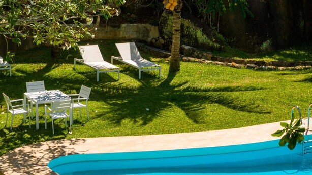 Piscina com borda curva, cercada por uma área gramada com espreguiçadeiras brancas e uma mesa.