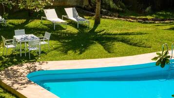 A swimming pool with a curved edge, surrounded by a grassy area with white lounge chairs and a table.