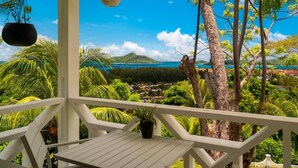 Varanda branca com vista para uma paisagem tropical, incluindo árvores e um corpo de água distante.