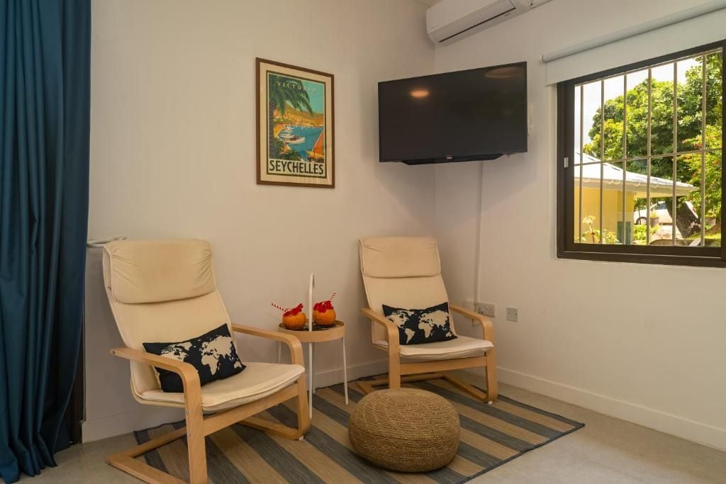 A room with two beige chairs with world map cushions, a small table with decorative items, a mounted TV, and a window with a view of greenery.