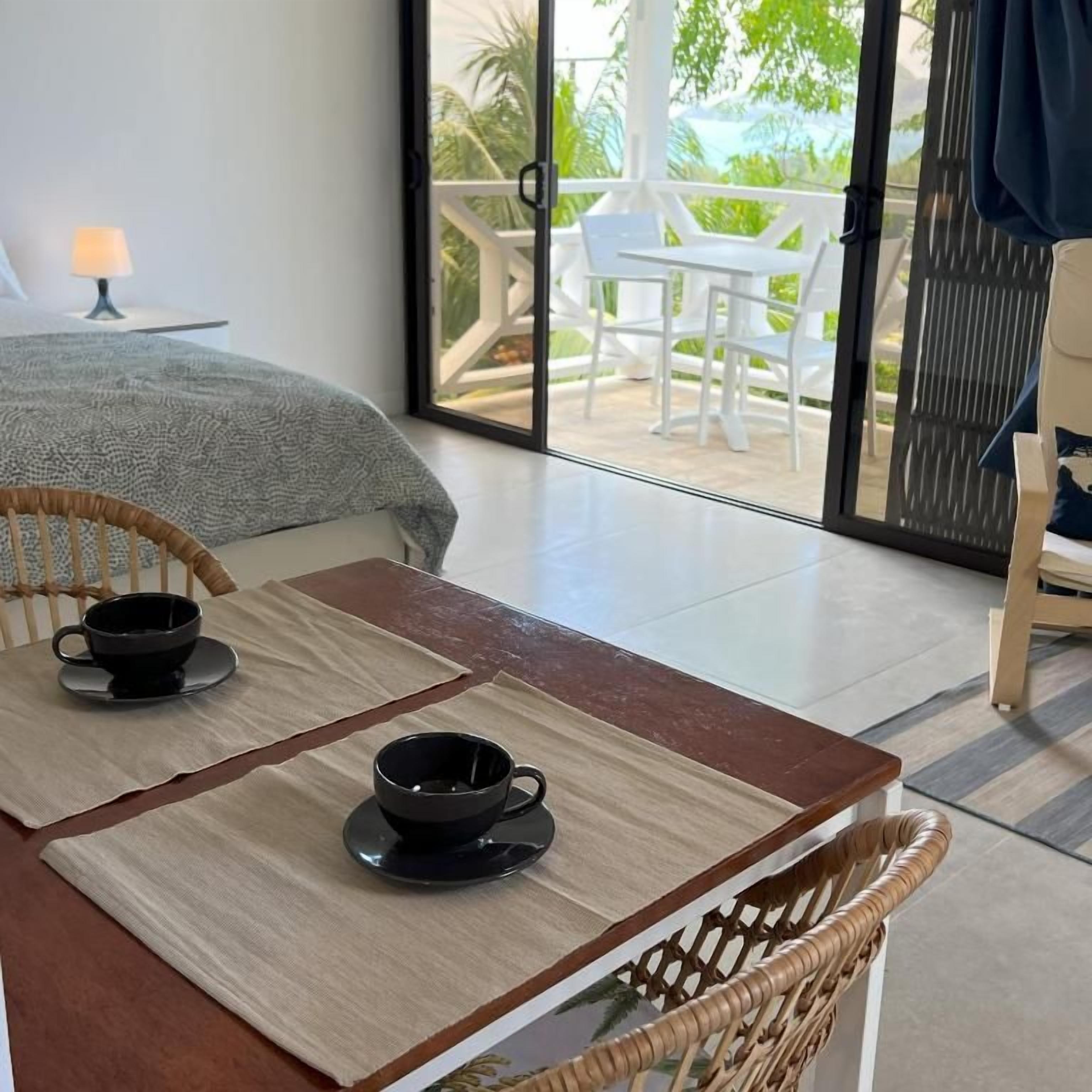 A room with a bed, a table with two cups, and a view of the outdoors through a glass door.